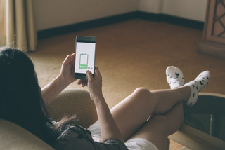 Symbolbild: Frau sitzt mit Handy auf dem Sofa, der Akku des Geräts ist leer