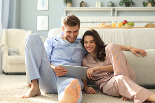 Junges Paar in Pyjamas sitzt auf dem Boden vor der Couch und blickt in einen Laptop Junges Paar in Pyjamas sitzt auf dem Boden vor der Couch und blickt in einen Laptop